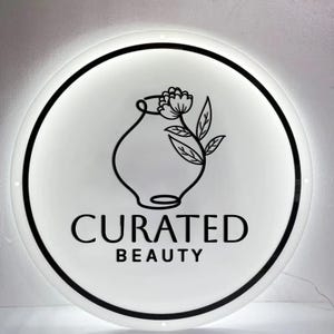 May include: A round, illuminated sign with a white face and a black border. The sign features a black line drawing of a vase with a flower and leaves. Below the illustration, the words "CURATED BEAUTY" are printed in black.