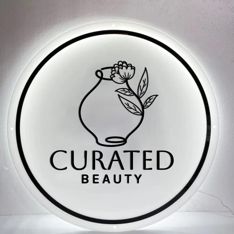 May include: A round, illuminated sign with a white face and a black border. The sign features a black line drawing of a vase with a flower and leaves. Below the illustration, the words "CURATED BEAUTY" are printed in black.