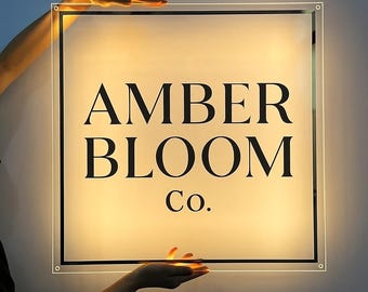 May include: A square, transparent acrylic sign featuring the black text AMBER BLOOM Co. centered on the surface. The sign is illuminated from behind, creating a soft, warm glow that highlights the lettering and the thin black border.