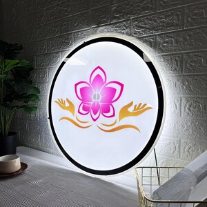 May include: Round, illuminated sign with a white background, featuring a pink flower design and gold-colored hands. The sign has a black border and is mounted on a wall. The overall theme is spa or wellness.