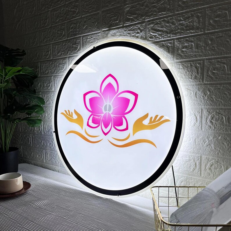 May include: Round, illuminated sign with a white background, featuring a pink flower design and gold-colored hands. The sign has a black border and is mounted on a wall. The overall theme is spa or wellness.