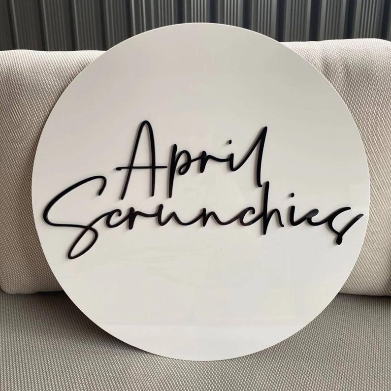 May include: A white, circular sign with the words "April Scrunchies" in black, cursive lettering. The sign is set against a neutral-toned background, suggesting a product display or shop sign.