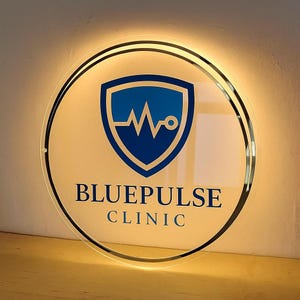 Op de afbeelding: Een rond, verlicht bord met de tekst "BLUEPULSE CLINIC". Het bord heeft een blauw schildlogo met een hartslagontwerp. Het heeft een zwarte rand en is van achteren verlicht.