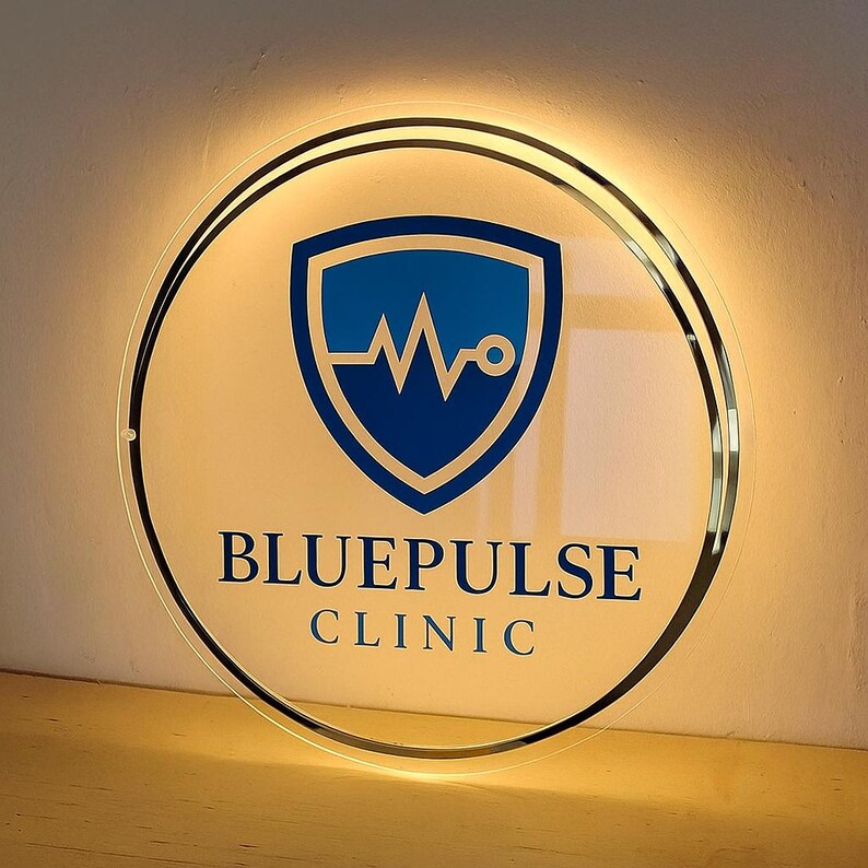 Op de afbeelding: Een rond, verlicht bord met de tekst "BLUEPULSE CLINIC". Het bord heeft een blauw schildlogo met een hartslagontwerp. Het heeft een zwarte rand en is van achteren verlicht.