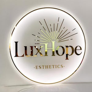 Puede incluir: Un letrero redondo iluminado con el texto dorado "LuxHope", un diseño de rayos de sol y la palabra "ESTHETICS" debajo. El letrero tiene un borde dorado y un fondo blanco, ideal para un negocio de estética.