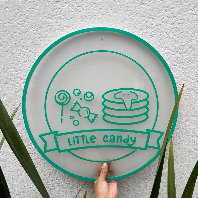May include: A round, clear sign with a teal border and teal graphics. The sign features illustrations of a lollipop, candy, and pancakes. The words "LiTTLE candy" are written in teal on a banner across the bottom.