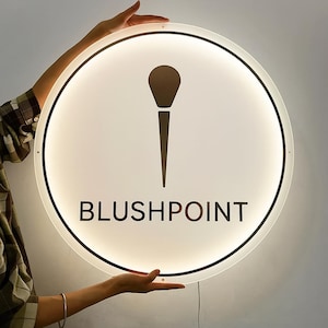 Puede incluir: Un letrero redondo e iluminado con una cara blanca y un borde negro. Presenta un icono de brocha de maquillaje y la palabra "BLUSHPOINT" en letras negras. El letrero est&aacute; siendo sostenido por una persona.