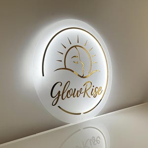 Puede incluir: Un letrero redondo e iluminado con fondo blanco y detalles dorados. El letrero presenta un dise&ntilde;o de sol y rostro, con las palabras "Glow Rise" en una escritura elegante. El letrero est&aacute; montado en una pared.