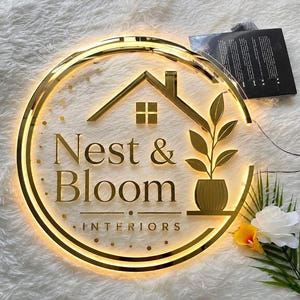 May include: A gold-colored, illuminated sign with the text "Nest & Bloom Interiors" in a circular design. The sign features a house silhouette, a potted plant, and a gold border. A black card is visible in the background.