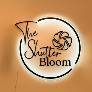 Puede incluir: Un letrero redondo e iluminado con las palabras "The Shutter Bloom" en una fuente elegante. El letrero presenta un borde negro y un gr&aacute;fico estilizado de lente de c&aacute;mara. El fondo es de un c&aacute;lido color naranja.