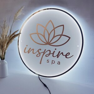 May include: A round, illuminated sign with a white background and a copper-colored lotus flower design. The sign reads "inspire spa" in a stylish font. The sign has a gold-colored trim and a black power cord.