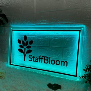 Op de afbeelding: Een rechthoekig bord met een zwarte rand en de tekst "StaffBloom" in het zwart. Het bord heeft een zwart bloemenontwerp en wordt verlicht met een turquoise gloed. Het bord is gemonteerd op een witte bakstenen muur.