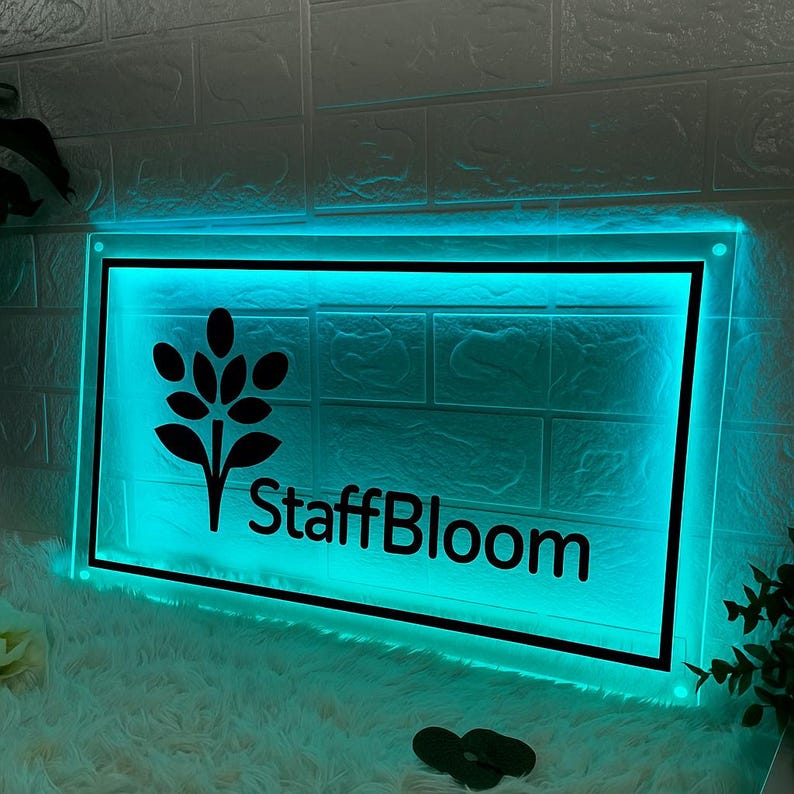 Op de afbeelding: Een rechthoekig bord met een zwarte rand en de tekst "StaffBloom" in het zwart. Het bord heeft een zwart bloemenontwerp en wordt verlicht met een turquoise gloed. Het bord is gemonteerd op een witte bakstenen muur.
