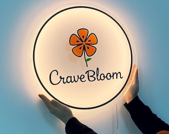 May include: A round, illuminated sign with the text "Crave Bloom" and a stylized orange flower. The sign is held by two hands against a light blue wall. The sign has a black border and a white background.