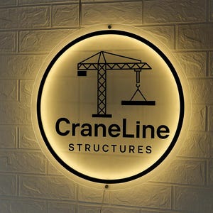 May include: A circular, backlit wall sign featuring a black graphic of a construction crane and the text CraneLine STRUCTURES in black lettering. The sign emits a warm, soft glow from behind its perimeter, highlighting the bold design.