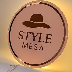 Op de afbeelding: Een ronde, ros&eacute;gouden plaat met de woorden "STYLE MESA" in donkerbruine letters. Een donkerbruine hoedafbeelding staat boven de tekst. Het bord heeft een reflecterende rand en is van achteren verlicht.