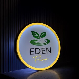 May include: A round sign with a yellow border and a light blue center. The sign features a green logo of leaves and water, with the words "EDEN Flow" in black and yellow script. The background is dark.