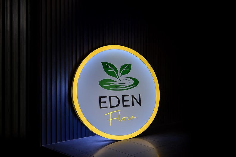 May include: A round sign with a yellow border and a light blue center. The sign features a green logo of leaves and water, with the words "EDEN Flow" in black and yellow script. The background is dark.