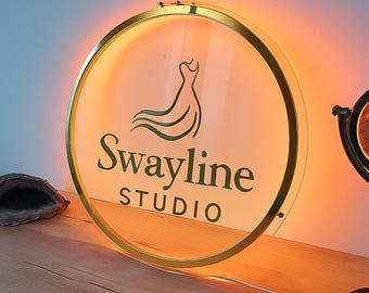 Puede incluir: Un letrero redondo e iluminado con un marco dorado. El letrero presenta el texto "Swayline STUDIO" en verde, junto con una silueta de vestido verde. El letrero está iluminado con un brillo cálido y anaranjado.