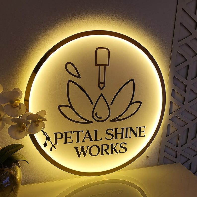 May include: A round, illuminated sign with a gold border. The sign features a black graphic of a lotus flower with a dropper and a water droplet. The text "PETAL SHINE WORKS" is below the graphic. The sign is lit with a warm, yellow glow.
