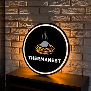 May include: A round, illuminated sign with a black background and white border. The sign features a nest design with an egg and the word "THERMANEST" in white. The sign is mounted on a dark brown surface against a brick wall.