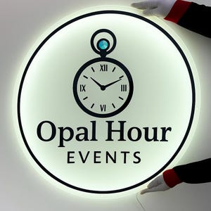 May include: A circular, backlit wall sign featuring a black graphic of a pocket watch with a blue gem accent at the top. The text Opal Hour EVENTS is printed in black in the center. The sign emits a soft white glow from its edges.