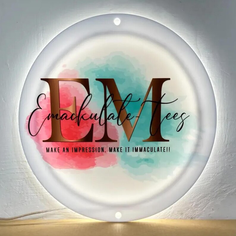 May include: A round, illuminated sign with the text "Emaculate Tees" in a copper-colored font. The background features watercolor splashes of red and teal. The sign also includes the text "MAKE AN IMPRESSION. MAKE IT IMMACULATE!!".