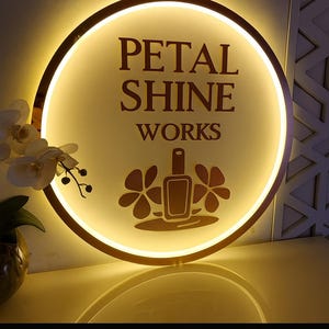 May include: A round, illuminated sign with the text "PETAL SHINE WORKS" in a serif font. The sign features a gold border and a central design of a nail polish bottle and flowers. The sign is lit with a warm, inviting glow.