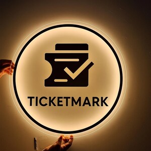 Puede incluir: Un letrero redondo e iluminado con un borde negro y una luz c&aacute;lida y brillante. El letrero presenta un icono de boleto negro con una marca de verificaci&oacute;n y la palabra "TICKETMARK" en letras negras en negrita.