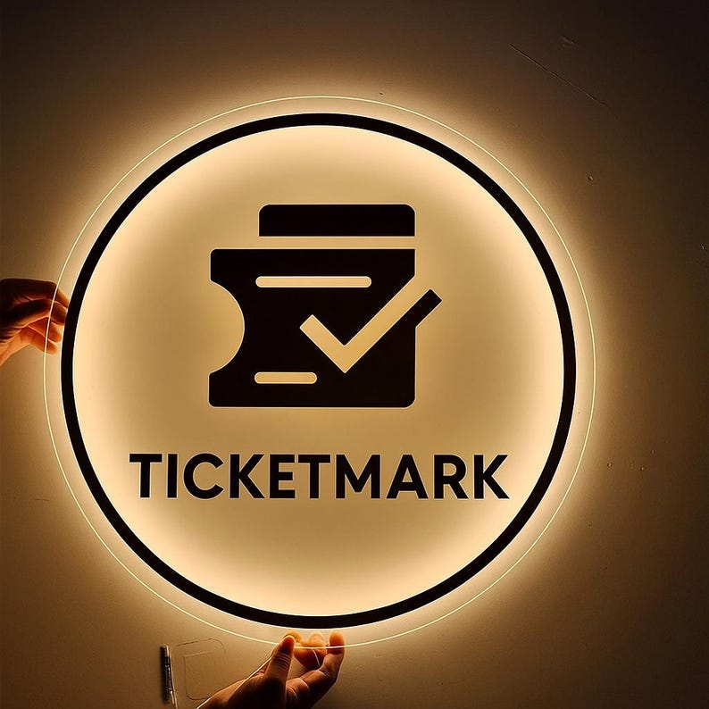 Puede incluir: Un letrero redondo e iluminado con un borde negro y una luz c&aacute;lida y brillante. El letrero presenta un icono de boleto negro con una marca de verificaci&oacute;n y la palabra "TICKETMARK" en letras negras en negrita.