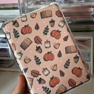 Può includere: Una custodia per telefono trasparente con un design autunnale. La custodia presenta uno sfondo rosa chiaro con illustrazioni di zucche, foglie, libri, ghiande, candele e tazze da caffè.