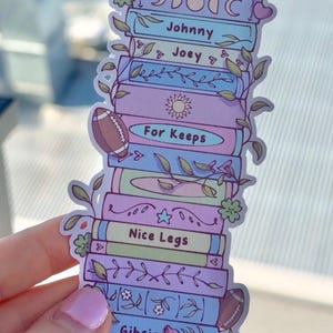 May include: A vertical sticker featuring a stack of pastel-colored books decorated with vines, flowers, and footballs. The spines display the text Aoife, Johnny, Joey, For Keeps, Nice Legs, Gibsie, Claire, and Shanon, along with various symbols.