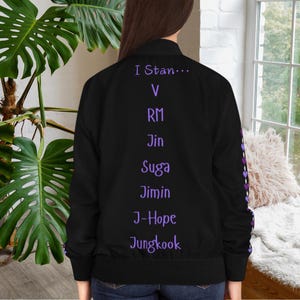 May include: Black bomber jacket with purple text that reads "I Stan..." followed by names. The jacket has a purple heart pattern on the sleeve. The jacket is shown from the back.