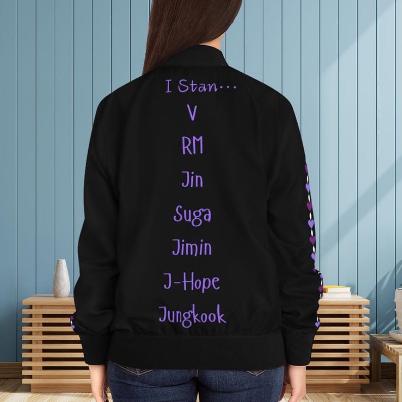 May include: Black bomber jacket with purple text that reads "I Stan..." followed by names. The sleeve has a purple heart design. The jacket is worn by a person.
