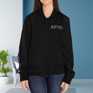 May include: Black bomber jacket with the word "ARMY" in purple script on the chest. The left sleeve features a pattern of purple hearts. The jacket has a zipper closure and two front pockets. The model is wearing blue jeans.