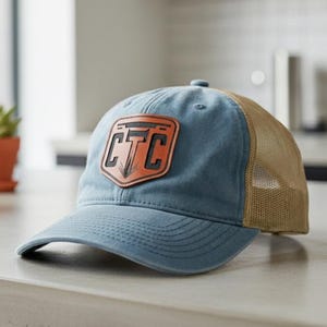 May include: A blue baseball cap with a tan mesh back and a leather patch. The patch features the letters "CTC" in a shield design. The hat is sitting on a light-colored countertop.