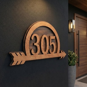 May include: Wooden address sign with the number 305, featuring an arrow design. The sign is made of light brown wood and mounted on a dark gray wall. The arrow points to the right, and the numbers are in a bold, stylized font.
