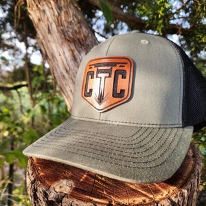 May include: A green and black trucker hat with a leather patch featuring the letters "CTC" in a shield design. The hat has a curved brim and a mesh back. The hat is sitting on a tree stump.