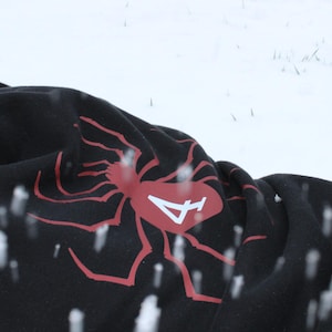 May include: Black hoodie with a red spider graphic on the front. The hoodie is lying on a snowy surface.