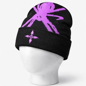 May include: A black knit beanie featuring a vibrant purple spider graphic on the crown and a small, purple four-pointed star symbol embroidered on the front cuff. The hat is displayed on a white mannequin head against a plain white background.