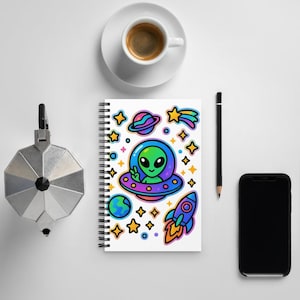 May include: A spiral-bound notebook featuring a colorful graphic print of a green alien in a blue and purple flying saucer, surrounded by planets, a rocket ship, and yellow stars on a white background, with a black pencil and coffee cup.