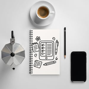 Könnte beinhalten: Ein weißes Spiralnotizbuch mit einer schwarzen Strichzeichnung eines Tablets mit Checkliste, zwei kleinen Notizblöcken, einem Stift, einem Bleistift und dekorativen Kritzeleien. Das Notizbuch liegt neben einer silbernen Kaffeemaschine, einer Tasse Kaffee, einem schwarzen Bleistift und einem schwarzen Smartphone.
