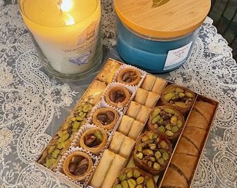 Puede incluir: Una caja cuadrada de pasteles baklava variados con capas de masa filo, pistachos picados y dulces enrollados rellenos de frutos secos, acompañada de una vela Yankee encendida y una vela azul Nature's Wick con tapa de madera.