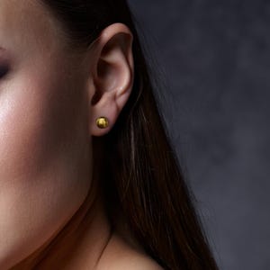 Pu&ograve; includere: Primo piano di un orecchio con un piccolo orecchino a bottone dorato. L'orecchino ha una forma rotonda e semplice. Lo sfondo &egrave; scuro e neutro, evidenziando l'orecchino, la pelle e i capelli.