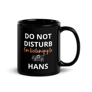 May include: Black coffee mug with white text that reads "Do Not Disturb I'm listening to HANS" with musical notes above the name.