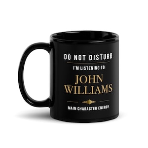 Pode incluir: Uma caneca de cerâmica preta com texto em branco e dourado que diz: DO NOT DISTURB I'M LISTENING TO JOHN WILLIAMS MAIN CHARACTER ENERGY, com um pequeno elemento gráfico dourado entre o nome e a última linha de texto.
