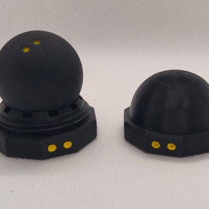 Puede incluir: Dos soportes de bolas de tenis de mesa negros con puntos amarillos. El soporte de la izquierda tiene una bola en la parte superior. Ambos soportes tienen una base hexagonal.