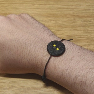 Könnte beinhalten: Ein schwarzes Armband mit einem runden schwarzen Anhänger mit drei gelben Punkten.