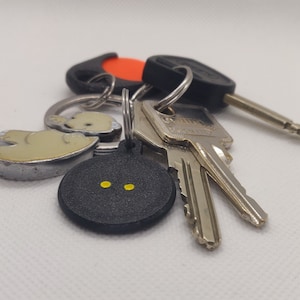Könnte beinhalten: Ein Schlüsselsatz mit einem schwarzen und silbernen Schlüsselanhänger, einem silbernen Schlüssel mit einem Bärenanhänger und einem silbernen Schlüssel mit einem schwarzen runden Anhänger mit zwei gelben Punkten.