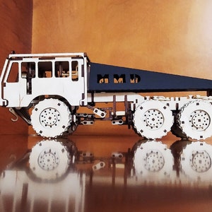 May include: A white wooden model truck with a black flatbed. The flatbed has the letters "MMD" cut out of it. The truck has four large wheels and a detailed chassis.